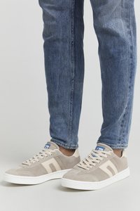 Beige Wildleder-Sneaker mit weißer Gummisohle, ausgestattet mit hellbeigen Streifen und einem blauen Etikett auf der Zunge. Getragen mit blauen Denim-Jeans.