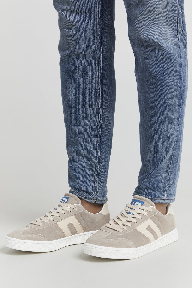 Beige Wildleder-Sneaker mit weißer Gummisohle, ausgestattet mit hellbeigen Streifen und einem blauen Etikett auf der Zunge. Getragen mit blauen Denim-Jeans.