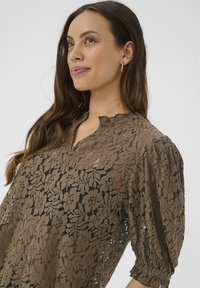 Lace bluse i mørkebrun med blomsterprint, pufærmer og splittet halsudskæring. Letvægts, tekstureret stof med semi-gennemsigtige detaljer.