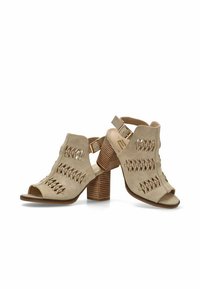 Sandalias de tacón abiertas en la parte delantera en color beige, con diseños recortados, correa con hebilla y un tacón de bloque de madera texturizada para mayor estabilidad. Material suave de ante.