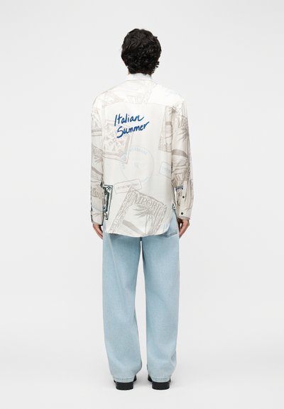 Homme aux cheveux bouclés portant une chemise blanche à manches longues avec le texte "Italian Summer" et des motifs, associée à un jean large bleu clair et des chaussures noires.