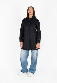 Cappotto nero fino al ginocchio con collo, bottoni frontali e tasche laterali; abbinato a jeans azzurri a gamba larga e sneakers bianche.