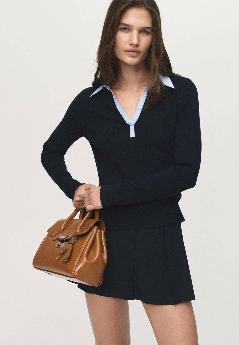 Woman wearing a black long-sleeve top with light blue collar and black skirt, holding a brown leather handbag with both hands.