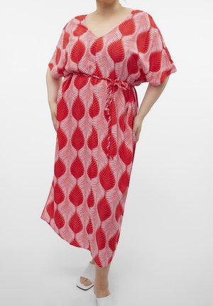 Kobieta w różowej sukience do połowy łydki z dużymi czerwonymi liściastymi wzorami, związaną w talii paskiem oraz białymi sandałami na obcasie z odkrytymi palcami.