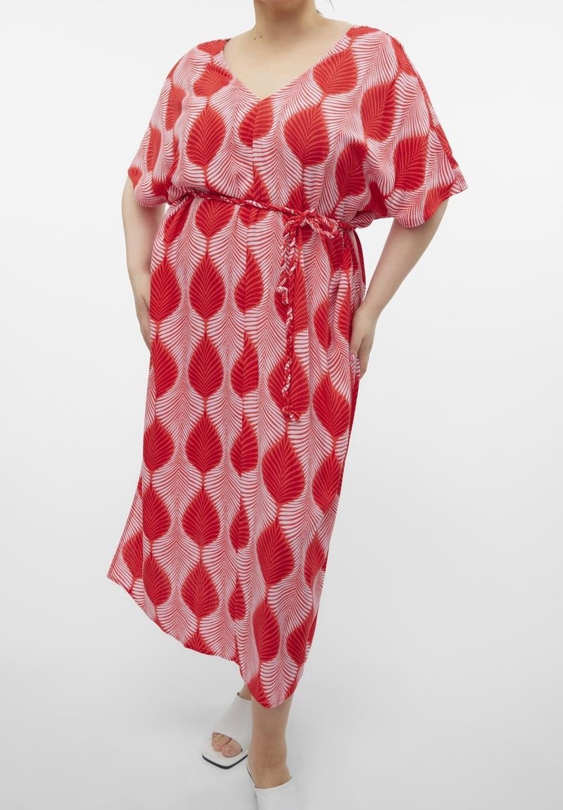 Femme portant une robe rose mi-longue avec de grands motifs de feuilles rouges, une ceinture nouée à la taille et des sandales blanches à talons ouvertes.