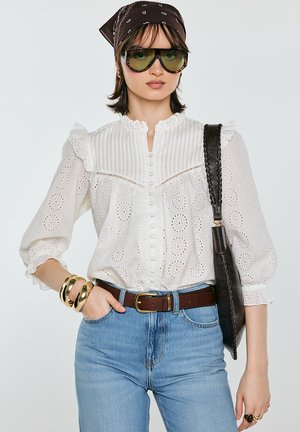 Kobieta ubrana w białą bluzkę z ażurowym wzorem, niebieskie dżinsy, brązowy pasek, duże złote bransolety, oversize’owe okulary przeciwsłoneczne, brązową chustę na głowę we wzory oraz czarną torbę na ramię.