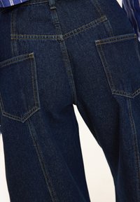 Pantalones vaqueros de pierna ancha en azul oscuro con dos bolsillos traseros y costuras en contraste. La tela tiene una textura suave y un diseño estructurado.