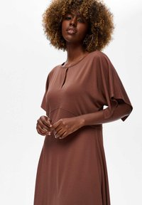 Vrouw met krullend haar, gekleed in grote oorringen en een bruine jurk met losse mouwen, staand tegen een eenvoudige witte achtergrond.