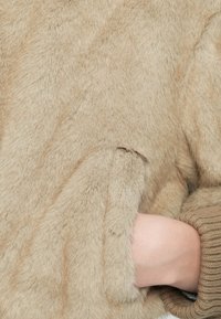 Tan umelá kožušina pulóver s plyšovou textúrou a rebrovými úpravami na rukávoch. Obsahuje bočnú kapsu viditeľnú na ľavej strane a mäkký, uvoľnený strih.