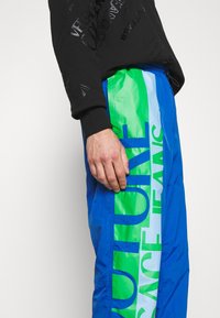 Modré a zelené větrové kalhoty s velkými nápisy "FUTURE" a "JEANS", hladkou texturou a lehkým designem, doplněné černou mikinou.