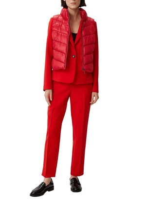 Vrouw in een rood pak met bijpassend mouwloos pufferjack, een zwarte top eronder en zwarte loafers, staand tegen een witte achtergrond.