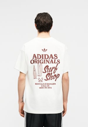 Άνδρας με την πλάτη γυρισμένη, φοράει λευκό μπλουζάκι Adidas με καφέ κείμενο: "Adidas Originals Surf Shop, Ενοικιάσεις & Επισκευές, Καλέστε μας 1800-3S-1972" και γραφικά με σανίδα σερφ.