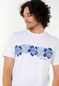 Hvid bomulds T-shirt med et blåt blomstertrykt over brystet, med "RIP CURL" tekst i blå under designet.
