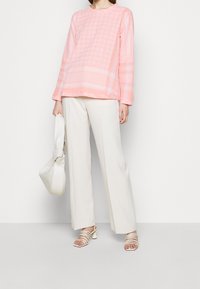 Różowa bluzka z długim rękawem w wzór w kratę houndstooth, zestawiona z luźnymi białymi spodniami i białymi sandałkami na obcasie, trzymająca białą torebkę.