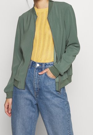 Frau trägt eine grüne Bomberjacke mit Reißverschluss über einem gelben Strickoberteil und hoch taillierte blaue Denimjeans mit Hand in der Tasche.