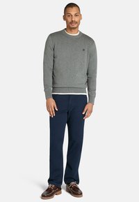 Grauer Sweatshirt mit Rundhalsausschnitt und gerippten Bündchen, über einem weißen Hemd getragen. Kombiniert mit dunkelblauen Hosen und braunen Schnürschuhen.