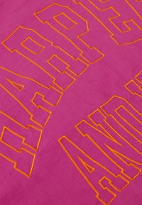 Roze stof met gewaagd oranje geborduurde tekst, met een gladde textuur en een vlakke lay-out. De letters hebben een dikke, blokachtige vorm.