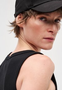 Fekete pamut ujjatlan felső széles pántokkal, texturált felülettel. A személy fekete sapkát visel, rövid, bozontos hajat mutatva.