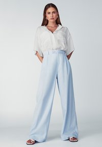 Svetlo modri široki hlače iz mehkega materiala, z gumbom za zapiranje in zankami za pas, v kombinaciji s prostornim belim srajcem.