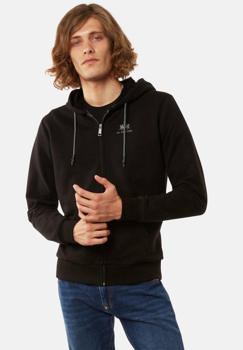 Sudadera con cremallera negra, hecha de una tela suave, que presenta una capucha con cordón, bolsillos frontales y un pequeño logo en el pecho. Llevada sobre una camiseta negra.