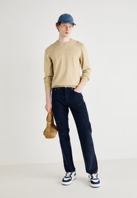 Sweatshirt beige à manches longues, pantalon bleu marine et baskets blanches avec accents marine ; tenant un sac à épaule beige. Fond uni.