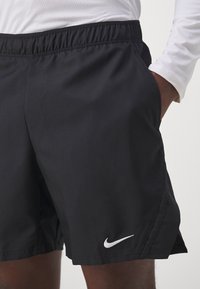 Černé sportovní šortky s elastickým pasem, bočními kapsami a bílým logem Nike na spodním levém lemu. Tkaninová textura.
