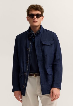 Junger Mann trägt eine dunkelblaue Jacke über einem marineblauen Pullover, beigefarbene Hose, geflochtener brauner Gürtel, dunkle Sonnenbrille und ein gemustertes Halstuch.