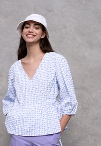 Top envolvente em azul claro com padrão, mangas três quartos, design peplum e um nó na cintura, combinado com um chapéu de balde branco.