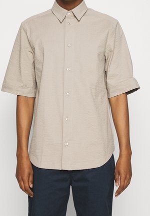 Personne portant une chemise beige à manches courtes avec un col pointu et un pantalon bleu marine foncé, debout devant un fond uni.
