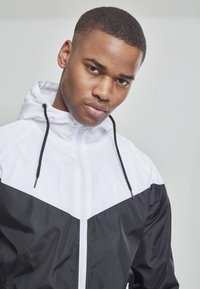 Chaqueta cortavientos ligera en blanco y negro, con cierre de cremallera en la parte delantera, capucha y un diseño geométrico contrastante con una textura suave.