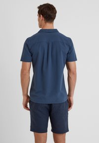 Short-sleeve shirt in textured dark blue fabric. Features a classic collar and back pleat. Worn with navy shorts.