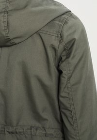 Olivgrön huvjacka med en slät yta, som har snedställda sömmar och elastisk nederkant för en figurnära passform. Hållbart tyg med minimal detaljering.