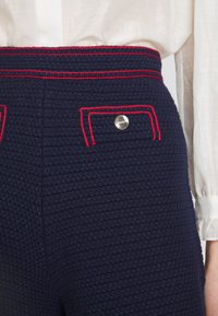 Pantalon bleu marine texturé taille haute avec des accents de couture rouges, bouton argenté sur la poche arrière, porté avec un chemisier blanc à manches longues.