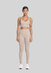 Žena koja stoji okrenuta prema naprijed, nosi bež sportski grudnjak, tajice visokog struka, bijele patike, s vidljivim tetovažama na rukama, na jednostavnoj sivoj pozadini.