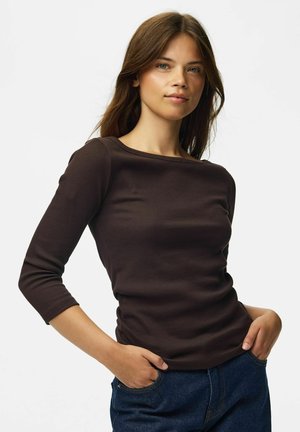 Jeune femme aux longs cheveux bruns portant un chemisier marron foncé ajusté à manches trois-quarts et un jean bleu, mains dans les poches, fond neutre.