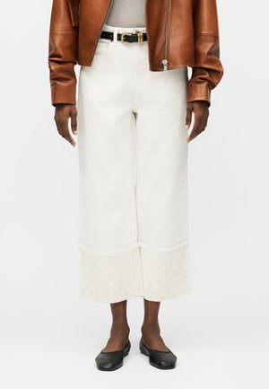 Άτομο που φοράει λευκά παντελόνια φαρδιά με κοντά μανίκια με μοτίβα, ένα καφέ δερμάτινο μπουφάν, μαύρη ζώνη και μαύρα μπαλαρίνες.