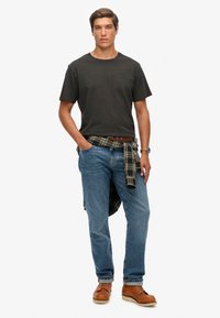 Czarna bawełniana koszulka, jasnoniebieskie dżinsy, koszula w kratę związana w talii, brązowe buty, zegarek na lewym nadgarstku. Fason slim-fit.