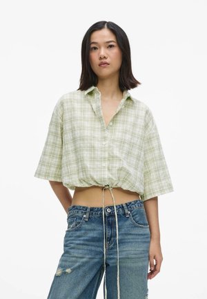 Donna che indossa una camicia corta a quadri verde chiaro con coulisse e jeans blu strappati a vita alta, in piedi su sfondo bianco.