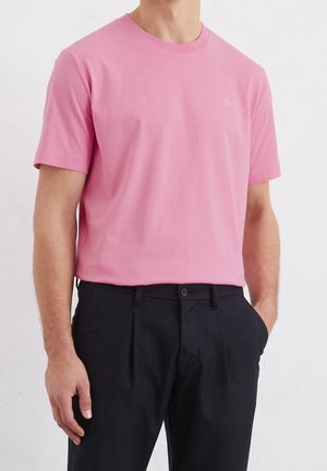 Lyserød bomulds t-shirt med korte ærmer, rund hals og et lille broderet logo på brystet. Kombineret med mørke bukser for kontrast.