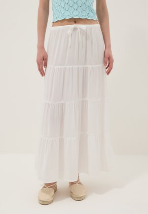 Vrouw draagt een gelaagde witte maxi rok met een aantrekkoord in de taille, een lichtblauw gehaakt crop top en beige espadrilles met enkelbandjes.