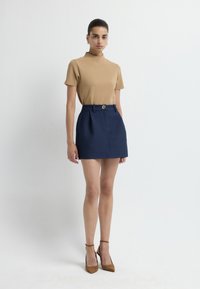 Beige aansluitend topje met korte mouwen, gecombineerd met een marineblauwe minirok met een ruitpatroon, voorzien van een voorplooi en een goudkleurige tailleknop. Bruine hakken.