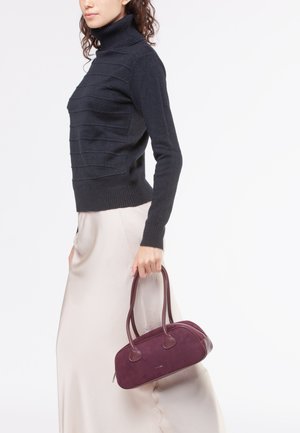 Borsa in suede bordeaux con dettagli in pelle liscia, forma arrotondata e doppi manici. Indossata con un dolcevita nero testurizzato e una gonna crema.