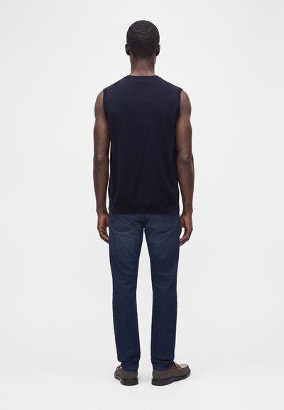 Homme debout de dos, portant un débardeur noir, un jean bleu foncé et des chaussures marron sur un fond blanc uni.