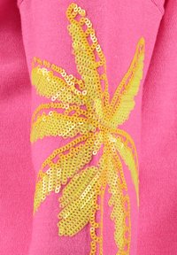 Tessuto di maglia rosa con un grande motivo di foglia di palma gialla in paillettes sulla manica. La consistenza è morbida, con un elemento decorativo leggermente in rilievo.