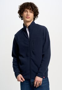 Tumesinine fliisi jakk kõrge kraega, esisepp ja rinnatasku. Omab siledat tekstuuri ja elastseid mansette. Kanduval valge T-särgi peal.