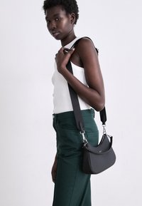 Sac à main noir en cuir lisse, au design courbé, avec des accessoires en argent, porté avec une bandoulière grise. Modèle en top blanc et pantalon vert.