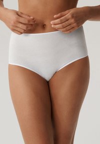 Kobieta poprawiająca gładką białą bieliznę w wysokim stanie, pokazująca tors, biodra i ręce na neutralnym tle.