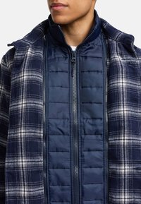 Veste à carreaux navy avec un intérieur matelassé. Caractéristiques d'une fermeture éclair, d'une capuche et de textures contrastantes entre la laine et le tissu rembourré.