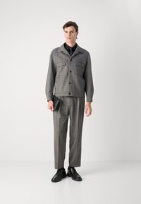Grau kariertes Jackett mit zwei Brusttaschen, kombiniert mit passenden karierten Hosen und schwarzen Schuhen. Hält eine schwarze Clutch.