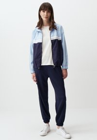 Giacca a vento blu chiaro e blu navy con striscia bianca, abbinata a joggers navy e sneakers bianche. Tessuto morbido, vestibilità casual, polsini elastici.